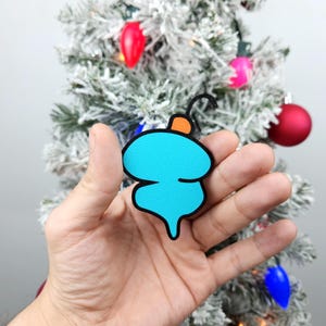 May include: A hand holding a blue and orange Christmas ornament shaped like a stylized Christmas tree. The ornament is made of felt and has a black outline.