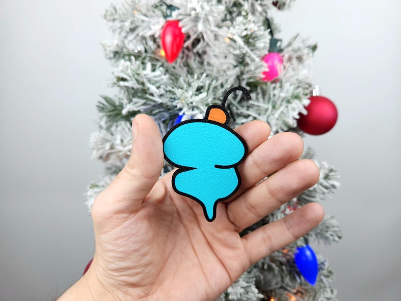 May include: A hand holding a blue and orange Christmas ornament shaped like a stylized Christmas tree. The ornament is made of felt and has a black outline.