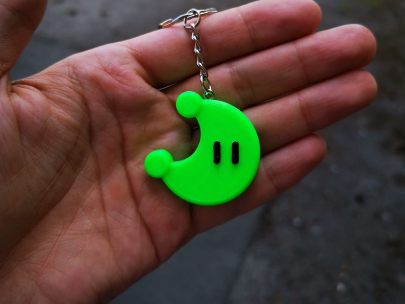 Puede incluir: Un llavero verde con forma de media luna con dos l&iacute;neas verticales negras en el centro.