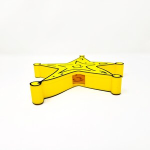 May include: A yellow star-shaped sheriff's badge with black outlines. The badge is made of plastic and has a hollow center.