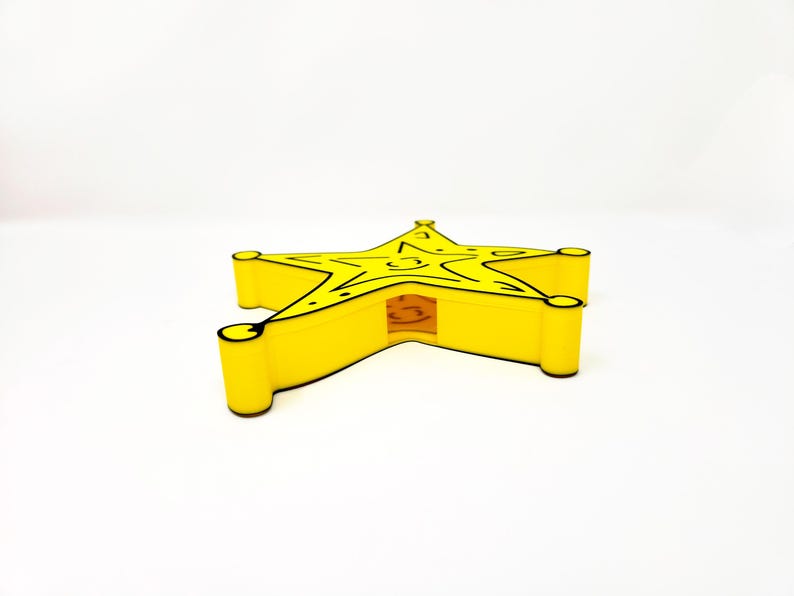 May include: A yellow star-shaped sheriff's badge with black outlines. The badge is made of plastic and has a hollow center.