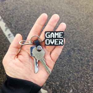 May include: A hand holding a set of keys with a black keychain that says "GAME OVER" in white pixelated letters.