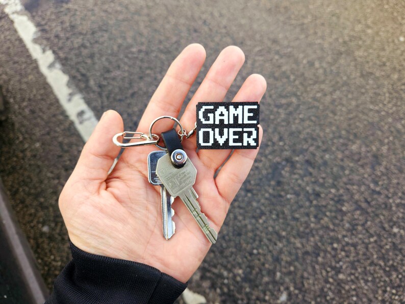 May include: A hand holding a set of keys with a black keychain that says "GAME OVER" in white pixelated letters.