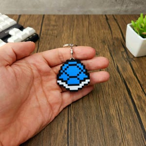 May include: A blue and white pixelated keychain shaped like a turtle shell. The keychain is being held in a hand.