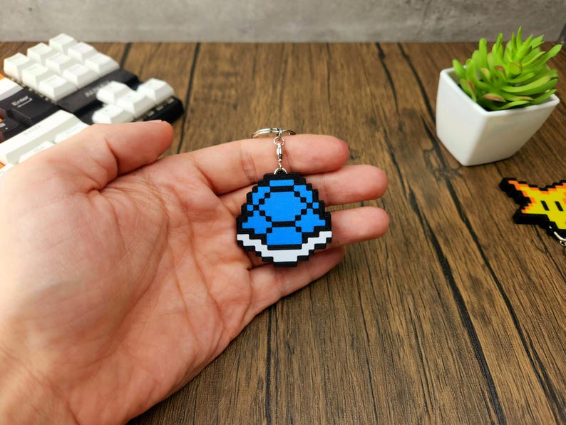 May include: A blue and white pixelated keychain shaped like a turtle shell. The keychain is being held in a hand.
