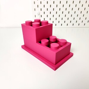 May include: A pink plastic organizer shaped like a stack of Lego bricks. The organizer has two levels, each with a row of circular holes for holding pens, pencils, or other small items.