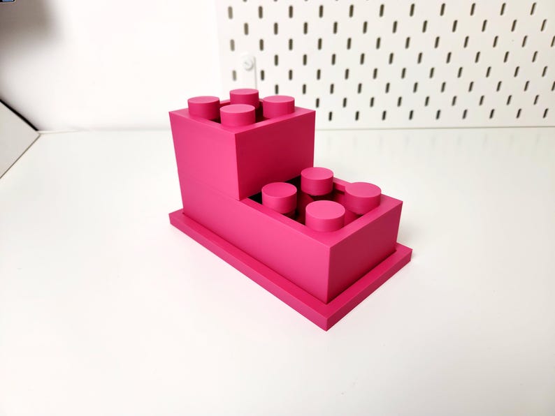 May include: A pink plastic organizer shaped like a stack of Lego bricks. The organizer has two levels, each with a row of circular holes for holding pens, pencils, or other small items.