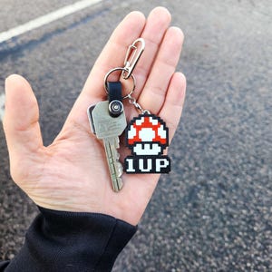 May include: A silver key with a black and silver keychain attached to a red and white pixelated mushroom charm with the text "1UP" on it.
