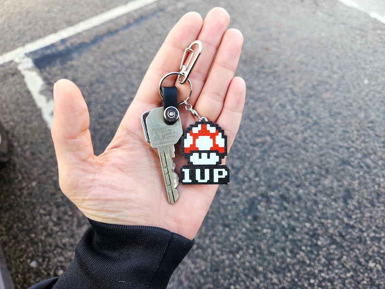 May include: A silver key with a black and silver keychain attached to a red and white pixelated mushroom charm with the text "1UP" on it.