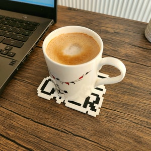 Puede incluir: Una taza de cer&aacute;mica blanca con un dise&ntilde;o negro y rojo se encuentra sobre un posavasos pixelado en blanco y negro. La taza est&aacute; llena de una bebida de caf&eacute; marr&oacute;n claro con un dise&ntilde;o de espuma blanca.