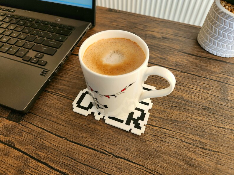 Puede incluir: Una taza de cer&aacute;mica blanca con un dise&ntilde;o negro y rojo se encuentra sobre un posavasos pixelado en blanco y negro. La taza est&aacute; llena de una bebida de caf&eacute; marr&oacute;n claro con un dise&ntilde;o de espuma blanca.