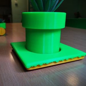 May include: A green plastic planter shaped like a Super Mario Bros. pipe with a small succulent plant growing inside. The planter has a yellow base with a scalloped edge.