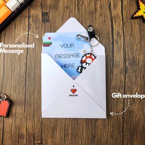 May include: A white envelope with a pixelated video game character on the front. The envelope has a red heart with the words "Open Me" printed on it. The envelope is on a wooden surface.