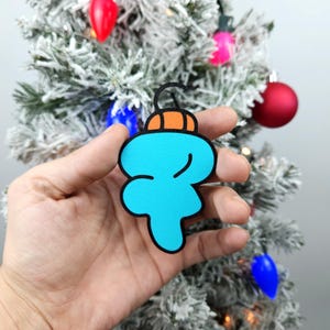 May include: A blue and orange Christmas ornament shaped like a cartoon character. The ornament is being held in a hand.