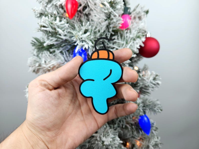 May include: A blue and orange Christmas ornament shaped like a cartoon character. The ornament is being held in a hand.