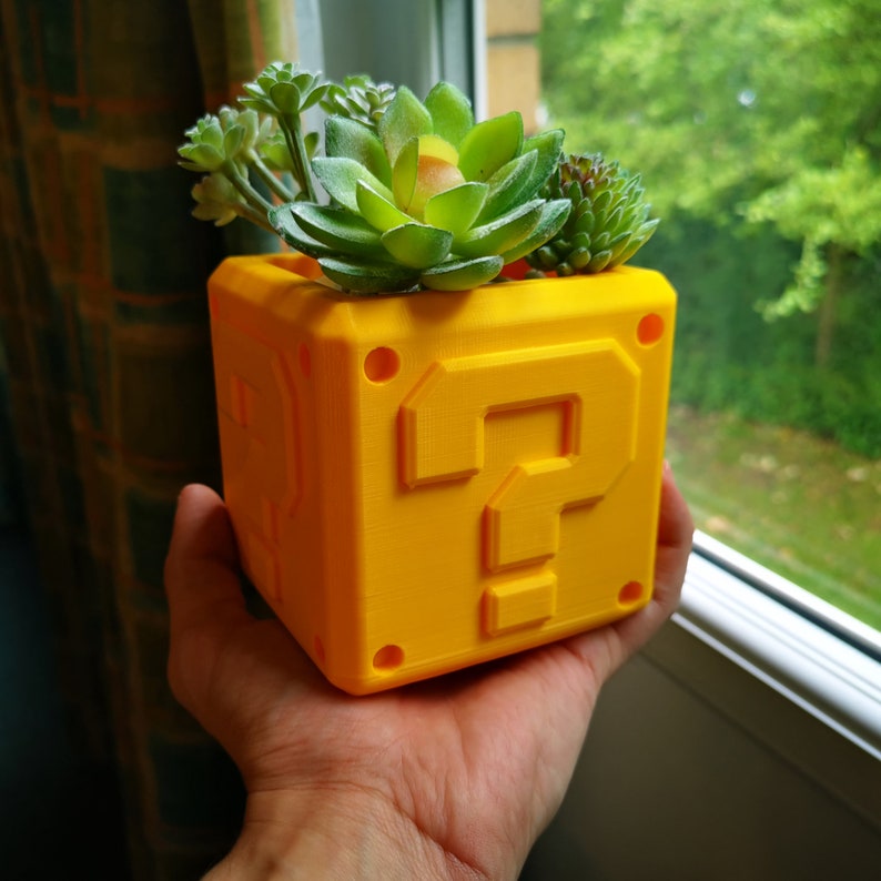 May include: A yellow question mark block planter with a variety of green succulents growing inside. The planter is made of 3D printed plastic and has a textured surface.