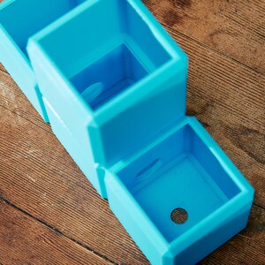 May include: Three stacked blue plastic square planters with drainage holes. The planters are empty and ready for planting.