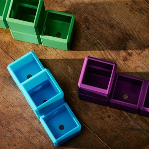May include: A selection of colourful, geometric planters in shades of green, blue, and purple. Each planter is a cube shape with a small drainage hole. The planters are arranged on a wooden surface.