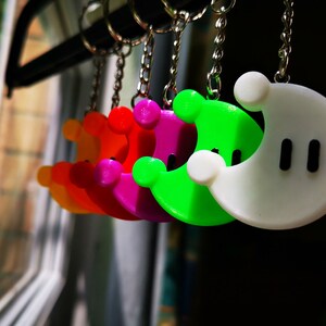 Puede incluir: Un conjunto de cinco llaveros coloridos con forma de luna creciente. Los llaveros son naranja, rojo, rosa, verde y blanco. Cada llavero tiene dos puntos negros en la luna creciente blanca.