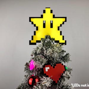 May include: A yellow pixelated star with a black outline sits atop a flocked Christmas tree. The star has a face with two black eyes. The tree is decorated with red and pink ornaments and lights. The text "LEDs not included" is visible at the bottom of the image.