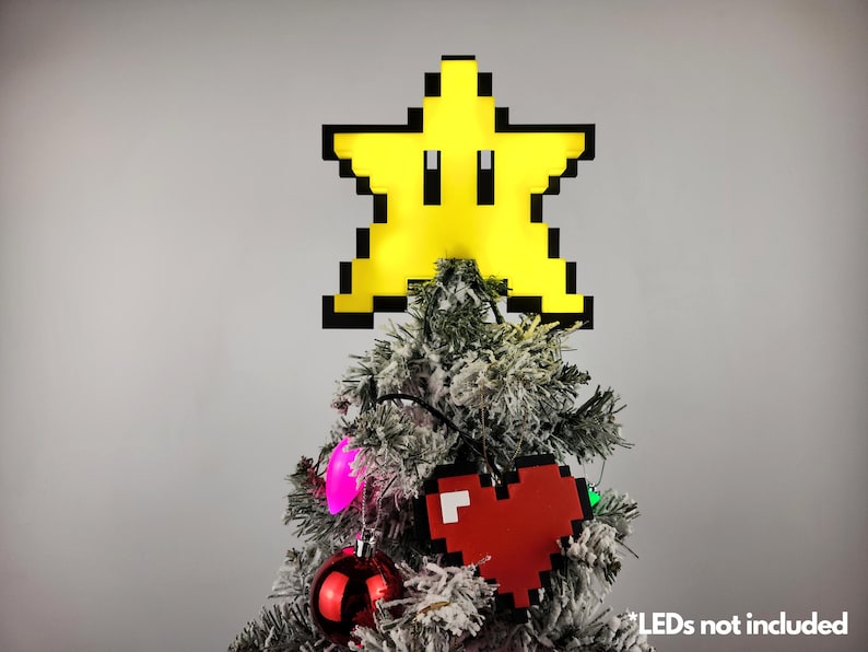 May include: A yellow pixelated star with a black outline sits atop a flocked Christmas tree. The star has a face with two black eyes. The tree is decorated with red and pink ornaments and lights. The text "LEDs not included" is visible at the bottom of the image.