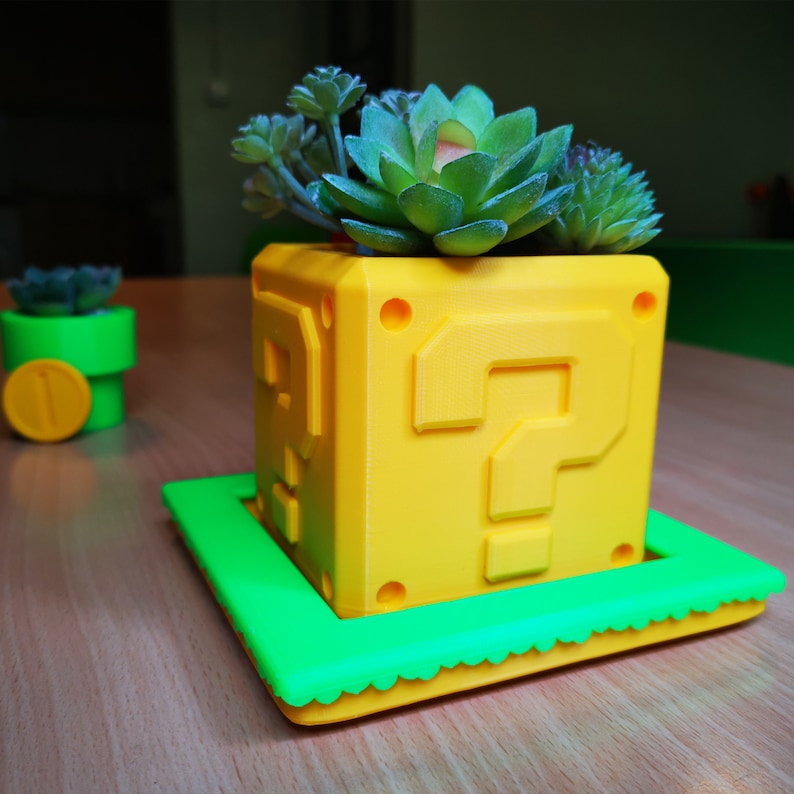 May include: A yellow question mark block planter with a green base. The planter is filled with a variety of succulents.