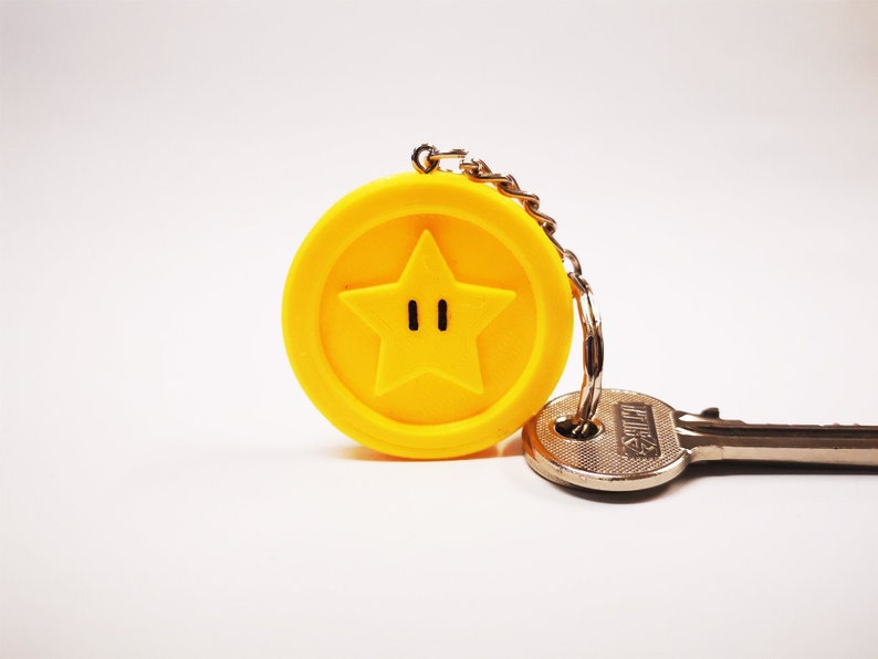 May include: A yellow keychain with a star-shaped charm featuring a black outline and two black eyes. The keychain is attached to a silver key.
