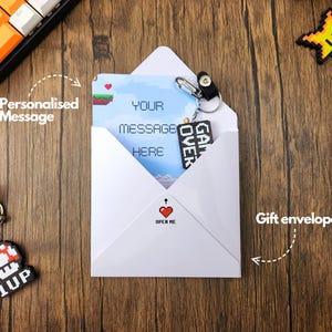 May include: A white envelope with a red heart and the text "Open Me" on the front. The envelope contains a card with a pixelated video game design and the text "Your message here". There are two pixelated video game keychains, one with a star and the other with a mushroom.