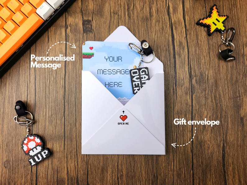 May include: A white envelope with a red heart and the text "Open Me" on the front. The envelope contains a card with a pixelated video game design and the text "Your message here". There are two pixelated video game keychains, one with a star and the other with a mushroom.