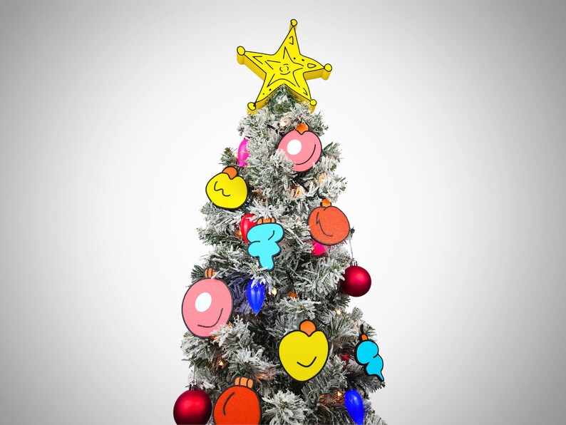 May include: A flocked artificial Christmas tree decorated with colorful cartoon ornaments, including a pink donut, a yellow smiley face, a red apple, and a blue cloud. A yellow star sits on top of the tree.