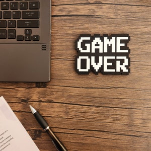 May include: A black laptop computer with a white keyboard is on a wooden table. A black and white pixelated sign that says "GAME OVER" is on the table. A silver pen is on the table next to a white piece of paper.