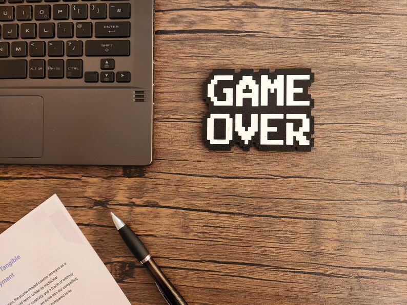May include: A black laptop computer with a white keyboard is on a wooden table. A black and white pixelated sign that says "GAME OVER" is on the table. A silver pen is on the table next to a white piece of paper.