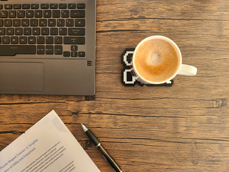 May include: A close-up of a wooden table with a laptop, a cup of coffee, a pen, and a piece of paper with the text "Puzzle Shaped Coaster: A Tangible" and "phor of Utility and Enjoyment".