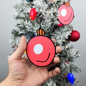 May include: A hand holds a red paper Christmas ornament with a white circle and a black smile. The ornament has a brown and orange top.