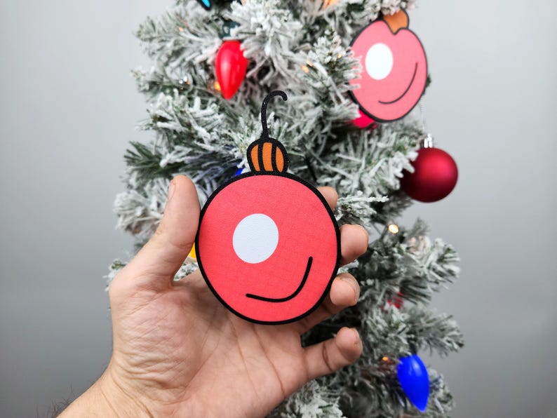 May include: A hand holds a red paper Christmas ornament with a white circle and a black smile. The ornament has a brown and orange top.
