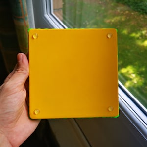 May include: A yellow square coaster with a green border and four clear rubber feet on the bottom.