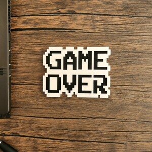 Puede incluir: Una pegatina blanca con texto pixelado negro que dice "GAME OVER" sobre una superficie de madera marr&oacute;n.