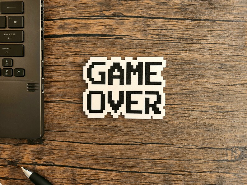 Puede incluir: Una pegatina blanca con texto pixelado negro que dice "GAME OVER" sobre una superficie de madera marr&oacute;n.