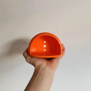 May include: Orange plastic half-dome with three small holes. The dome is held in a person's hand.