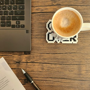 Puede incluir: Una taza de cer&aacute;mica blanca con una bebida de caf&eacute; marr&oacute;n en su interior, sentada sobre un posavasos blanco con texto pixelado negro que dice "Coffee". La taza est&aacute; sobre una mesa de madera con un port&aacute;til y un bol&iacute;grafo.
