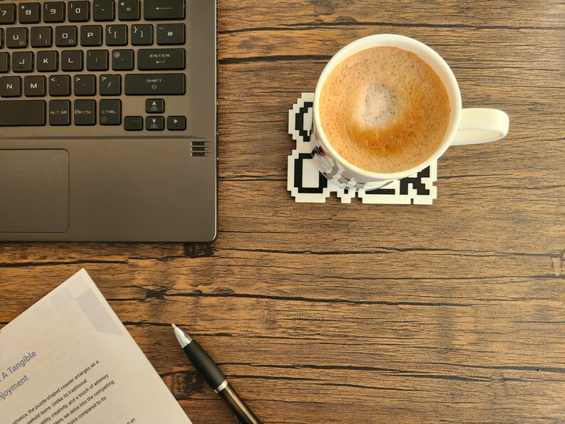 Puede incluir: Una taza de cer&aacute;mica blanca con una bebida de caf&eacute; marr&oacute;n en su interior, sentada sobre un posavasos blanco con texto pixelado negro que dice "Coffee". La taza est&aacute; sobre una mesa de madera con un port&aacute;til y un bol&iacute;grafo.