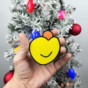May include: A yellow Christmas ornament shaped like a heart with a smiling face and a small orange ornament on top.