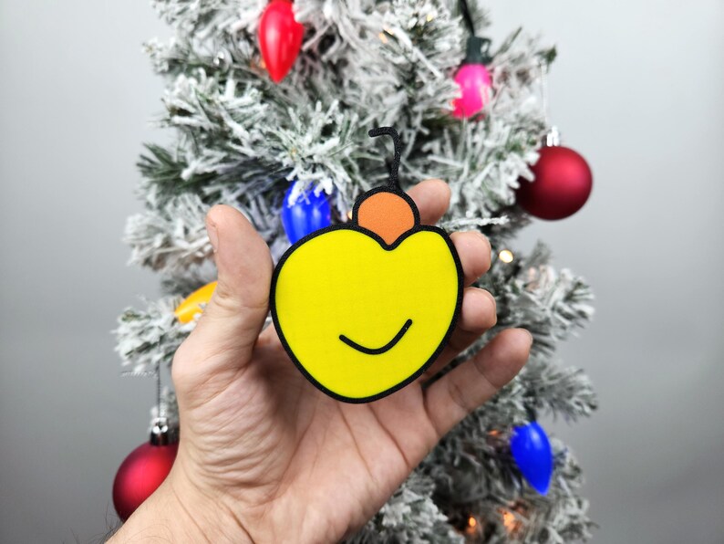 May include: A yellow Christmas ornament shaped like a heart with a smiling face and a small orange ornament on top.