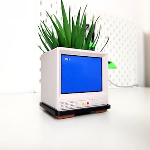 Pu&ograve; includere: Un vaso da fiori bianco in stile retr&ograve; a forma di televisore con uno schermo blu e una pianta verde che cresce dalla parte superiore. Il vaso ha una base nera e una piccola griglia dell'altoparlante sul davanti. Il testo "RV1" &egrave; visibile sullo schermo.