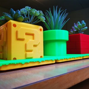 May include: Three 3D printed planters shaped like iconic Super Mario Bros. items. A yellow question block planter with a green base, a green pipe planter, and a red brick planter. Each planter has a small succulent plant inside.