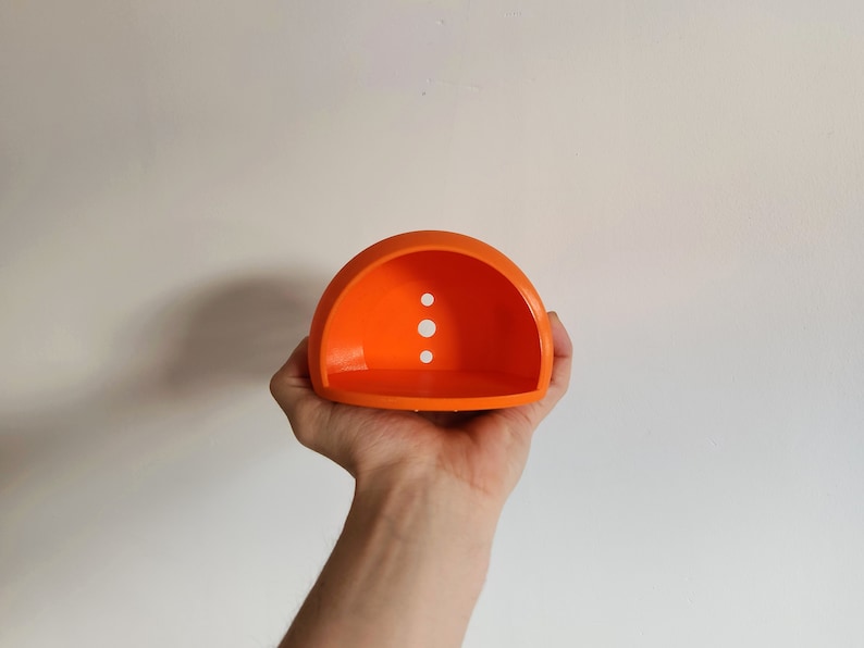 Op de afbeelding: Oranje plastic halve koepel met drie kleine gaten. De koepel wordt in de hand van een persoon gehouden.