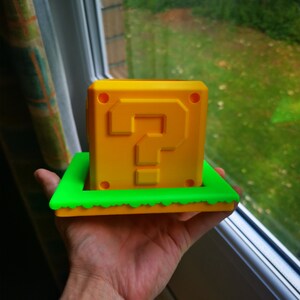 May include: A yellow 3D printed question mark block with a green base. The block is a popular item from the Super Mario Bros. video game series.