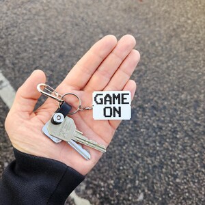 May include: A silver key chain with two keys and a white tag that says "GAME ON" in black pixelated letters.