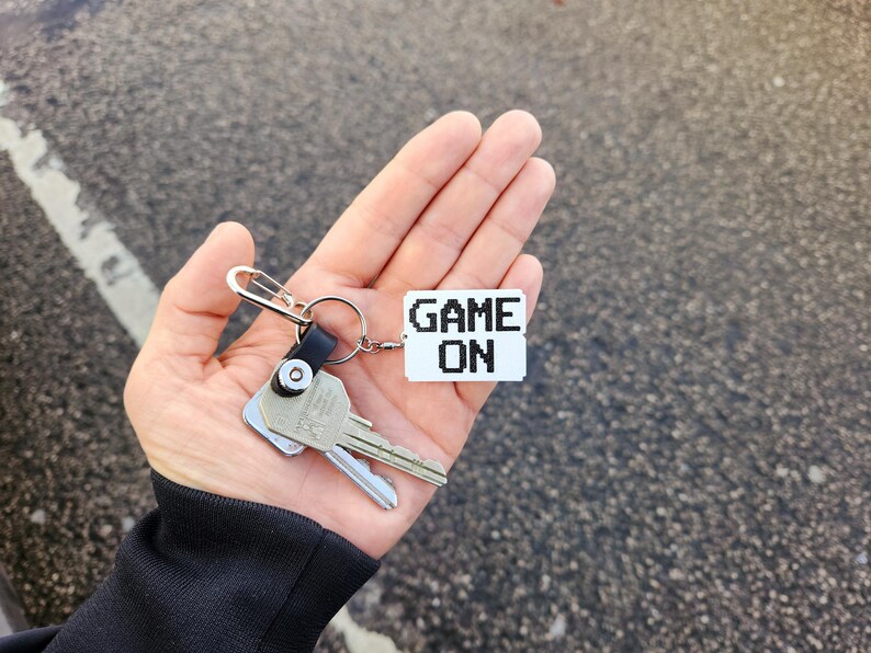 May include: A silver key chain with two keys and a white tag that says "GAME ON" in black pixelated letters.