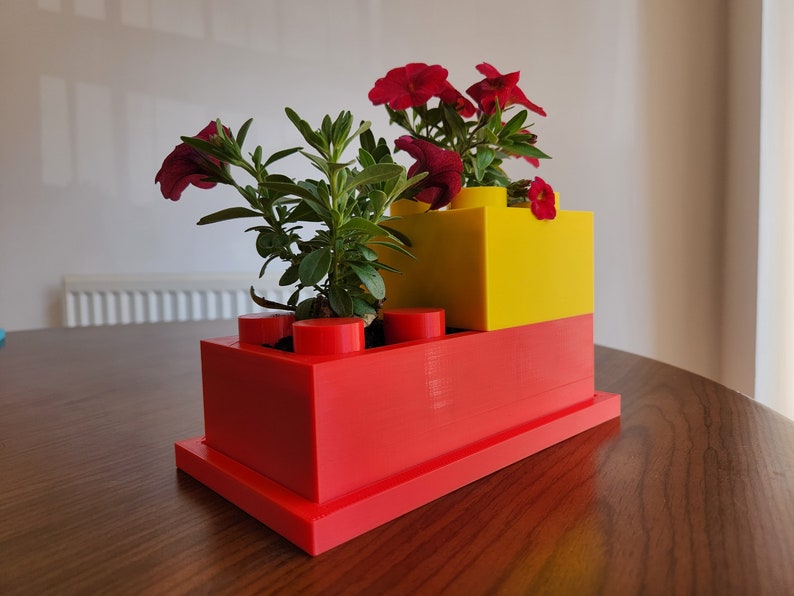 May include: A red and yellow Lego-style planter with a red base and a yellow top. The planter is filled with green plants and red flowers.